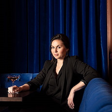 Beverage Director Amy Racine sitting in an upscale bar with dark blue velvet curtains.