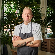 Executive Chef Warren Baird standing in an upscale setting in a white chef’s coat and apron.